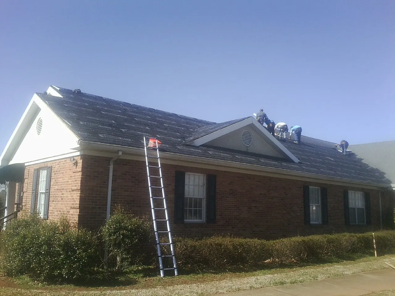 Crew tearing shingles off a brick ranch roof with a ladder set — Emergency Roof Repair work by our crew in Wellborn