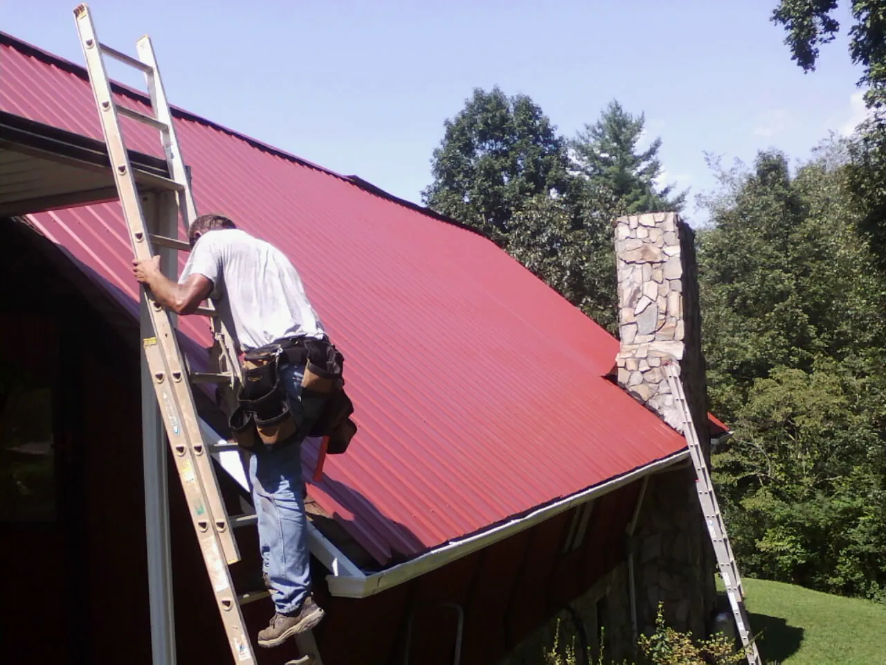 Roofer on a ladder beside a red metal roof and stone chimney — Soffit & Fascia Repair by Roofing Brothers in Wellborn